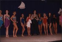 Ten Stevens models stand in a line in one-piece swimsuits with a male model in fishing gear during the Boat Show Fashion Show