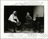 Jay McShann playing piano with unidentified man holding a tambourine